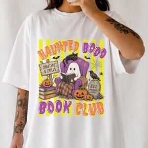 Könnte beinhalten: Ein weißes T-Shirt mit einem Grafikdruck mit gelben vertikalen Streifen und dem Text "HAUNTED BOOO BOOK CLUB". Das Design zeigt ein Gespenst, das in einem violetten Sessel ein Buch liest, eine schwarze Katze, Kürbisse, einen Grabstein und ein Schild mit der Aufschrift "CHAPTERS & CHILLS".