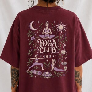 May include: A maroon oversized t-shirt featuring a graphic on the back with the text YOGA CLUB. The design includes illustrations of people in yoga poses, a crescent moon, a sun, flowers, a tea cup, a bowl, and a rolled-up yoga mat in purple.