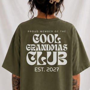 Op de afbeelding: Een olijfgroen t-shirt met witte tekst op de achterkant die luidt PROUD MEMBER OF THE COOL GRANDMAS CLUB EST. 2027. Het shirt heeft korte mouwen en een losse pasvorm, met de afbeelding gecentreerd op het bovenste gedeelte van de rug.