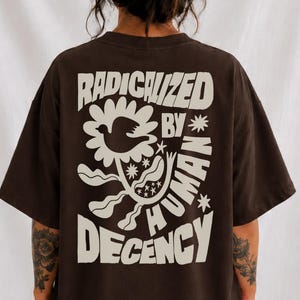 Puede incluir: Una camiseta de manga corta de color marrón oscuro con un gran gráfico de color crema en la espalda. El diseño incluye una flor estilizada con un pájaro en el centro, rodeada de estrellas y el texto RADICALIZED BY HUMAN DECENCY en negrita.