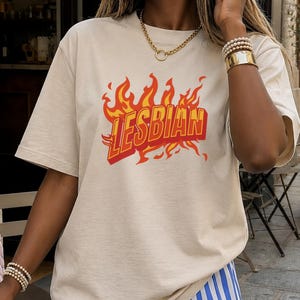 Könnte beinhalten: Ein sandfarbenes T-Shirt mit dem Wort "LESBIAN" in roten und gelben Flammen. Die Person trägt blau-weiß gestreifte Shorts. Eine rosa Handtasche und ein Getränk mit Strohhalm stehen auf einem Tisch.