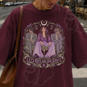 Peut inclure: Un t-shirt oversize marron avec une illustration graphique de trois personnages assis sur un banc, entourés de vignes florales et d'un croissant de lune. Le texte "LET'S RECLAIM OUR SPACE" est imprimé dans une police décorative en bas du motif.