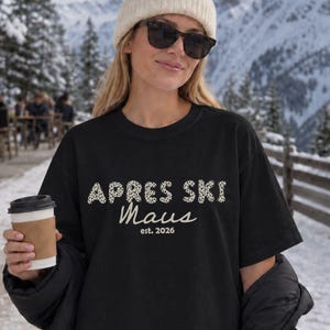 Könnte beinhalten: Ein schwarzes kurzärmeliges T-Shirt mit dem Text "APRES SKI" in einem weißen Leopardenmuster, gefolgt von dem Wort "Maus" in weißer Schreibschrift und "est. 2026" in einer kleineren weißen Schrift, getragen mit einer beigen Mütze und schwarzer Jacke.