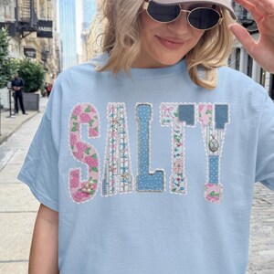 Könnte beinhalten: Hellblaues T-Shirt mit dem Wort "SALTY" in einem Patchwork-Design. Die Buchstaben sind mit floralen und gemusterten Stoffen in Rosa, Blau und Weiß verziert. Das Shirt hat kurze Ärmel und einen Rundhalsausschnitt.