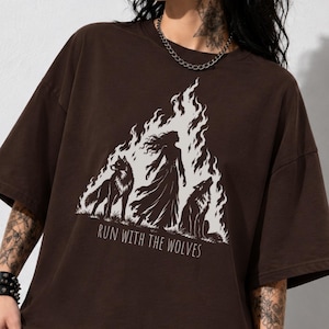 May include: A dark chocolate brown oversized t-shirt featuring a white graphic print of a person standing between two wolves amidst flames. The text "RUN WITH THE WOLVES" is printed below the illustration in a simple, clean font.
