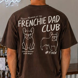 Könnte beinhalten: Ein dunkelbraunes Kurzarm-T-Shirt mit weißem Text, der Proud Member of the FRENCHIE DAD CLUB, Life is better with dogs! und #FINN lautet, ergänzt durch Strichzeichnungen von zwei Französischen Bulldoggen auf der Rückseite.