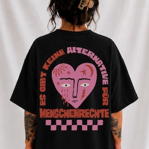 Könnte beinhalten: Ein schwarzes T-Shirt mit einem Rückendruck, der ein rosafarbenes herzförmiges Gesicht zeigt, umgeben von dem orangefarbenen Text "ES GIBT KEINE ALTERNATIVE FÜR MENSCHENRECHTE" und einem rosa-schwarzen Schachbrettmuster am unteren Rand des Motivs.