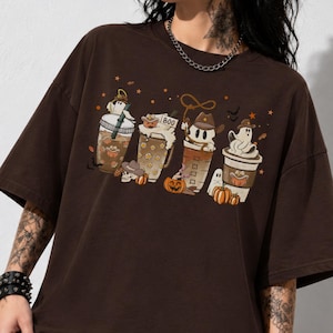 Pode incluir: Uma t-shirt de manga curta castanho-escura com uma impressão gráfica de quatro chávenas de café decoradas com fantasmas, abóboras e chapéus de cowboy. O design central inclui pequenas estrelas e o texto BOO numa das chávenas.