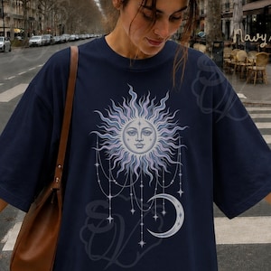 Könnte beinhalten: Ein marineblaues T-Shirt mit einem Grafikdruck einer stilisierten Sonne mit menschlichem Gesicht, umgeben von strahlenden Lichtstrahlen, hängenden Sternanhängern und einer Mondsichel. Das Shirt hat kurze Ärmel und einen Rundhalsausschnitt.