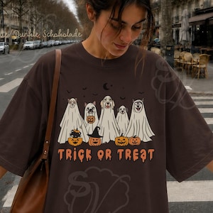 Könnte beinhalten: Ein dunkelbraunes Kurzarm-T-Shirt mit einer Grafik von fünf Hunden, die als Geister in weißen Laken verkleidet sind. Unter den Hunden befinden sich orangefarbene Kürbisse und der Schriftzug "TRICK OR TREAT" in tropfenden orangefarbenen Buchstaben.