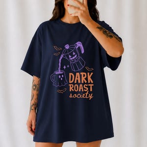 May include: A navy blue short-sleeve t-shirt featuring a purple graphic of a moka pot pouring liquid into a ghost-shaped mug. Small orange bats surround the design, and the text "DARK ROAST society" is printed in orange lettering below the mug.