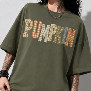 Könnte beinhalten: Ein olivgrünes Kurzarm-T-Shirt mit dem Wort PUMPKIN auf der Brust. Jeder Buchstabe ist mit einem anderen Muster gefüllt, darunter orange-weißes Vichy-Karo, florale Drucke und herbstliche Motive in verschiedenen Braun- und Beigetönen.