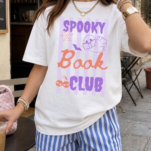 Könnte beinhalten: Weißes T-Shirt mit dem Text "SPOOKY Book CLUB" in Lila und Orange, mit einer Spinnennetz-, Geister- und Totenkopf-Grafik. Das Model trägt blau-weiß gestreifte Shorts. Das Gesamtthema ist Halloween und Buchclub.