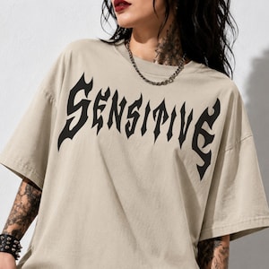 Könnte beinhalten: Ein beiges Kurzarm-T-Shirt mit dem Wort SENSITIVE, das in einer großen, schwarzen Schriftart im Gothic-Stil auf die Brust gedruckt ist. Das Kleidungsstück hat einen Rundhalsausschnitt und eine lockere Passform mit überschnittenen Schultern.