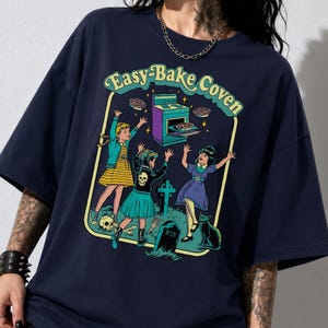 May include: A navy blue short-sleeve t-shirt featuring a graphic print with the text "Easy-Bake Coven". The illustration depicts three figures in a graveyard setting with a purple oven, a black cat, and tombstones, all framed in a light yellow border.