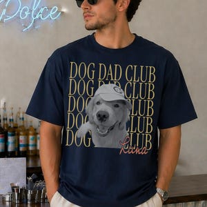 May include: A navy blue short-sleeve t-shirt featuring a repeating gold text pattern that reads DOG DAD CLUB. Centered on the chest is a black and white photograph of a dog wearing a baseball cap, with the name Luna written in cursive below.