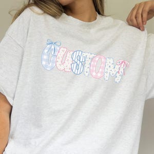 Könnte beinhalten: Ein hellgraues, übergroßes T-Shirt mit dem Wort "CUSTOM" in einem pastellfarbenen Farbblock-Design. Die Buchstaben sind mit Vichy-, Blumen- und Sternmustern sowie Schleifen verziert. Das Shirt wird mit blau-weiß gestreiften Shorts kombiniert.