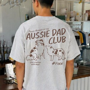 May include: A light grey short-sleeve t-shirt featuring brown text and illustrations on the back. The text reads PROUD MEMBER OF THE AUSSIE DAD CLUB, Life is better with dogs!, and #FINN. Two brown and white dog illustrations are centred.