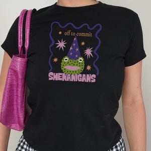 Könnte beinhalten: Schwarzes T-Shirt mit einem skurrilen Frosch-Motiv, der einen Zaubererhut trägt, umgeben von Sternen und den Worten "off to commit shenanigans". Das Shirt hat einen klassischen Rundhalsausschnitt.