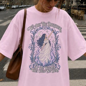 Könnte beinhalten: Ein hellrosa T-Shirt mit einer grafischen Illustration einer Figur mit langem dunklem Haar, die nach einer Mondsichel greift, umgeben von floralen Motiven und Flammen. Der Text lautet, We Are The Daughters Of The Witches They Could Not Burn.