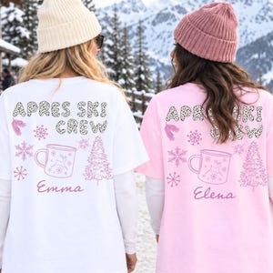 Könnte beinhalten: Zwei T-Shirts, eines in Weiß und eines in Hellrosa, mit dem Schriftzug APRES SKI CREW in Leopardenmuster. Die Designs zeigen rosa Schneeflocken, einen Tannenbaum und eine Tasse, mit den Namen Emma und Elena darunter gedruckt.