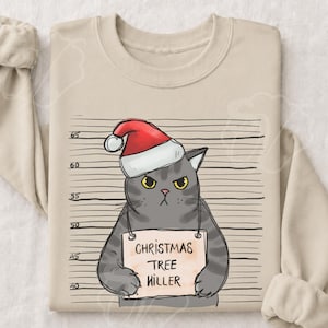 Könnte beinhalten: Ein beigefarbenes Langarm-Sweatshirt mit einer Grafik einer grauen getigerten Katze, die eine rot-weiße Weihnachtsmütze trägt. Die Katze ist vor einem Hintergrund mit einer Messskala abgebildet und hält ein Schild mit der Aufschrift CHRISTMAS TREE KILLER.
