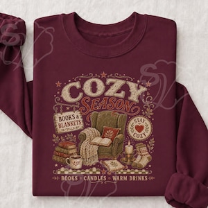 Puede incluir: Una sudadera de manga larga color granate con un estampado gráfico que incluye el texto "COZY SEASON" y "BOOKS & BLANKETS". La ilustración muestra un sillón con una manta, una pila de libros, una taza, una vela y calcetines con estrellas.