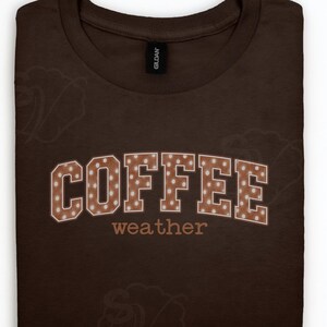 Op de afbeelding: Een donkerbruin t-shirt met ronde hals met het woord COFFEE in grote blokletters met een wit stippenpatroon, geplaatst boven het woord weather in een kleiner, lichtbruin cursief lettertype. Het shirt heeft een Gildan-merklabel.