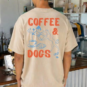 Może przedstawiać: Beżowa koszulka z napisem "COFFEE & DOGS" w kolorze pomarańczowym i niebieskim. Projekt zawiera ekspres do kawy, filiżankę, rogalik i sylwetkę psa. Koszulka ma luźny, obszerny krój.
