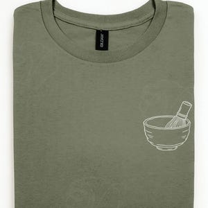 Może przedstawiać: Oliwkowy t-shirt z okrągłym dekoltem i białą grafiką liniową po prawej stronie. Grafika przedstawia miseczkę do matchy z bambusową trzepaczką w środku. Koszulka posiada czarną metkę na karku z nadrukowaną na biało nazwą marki Gildan.