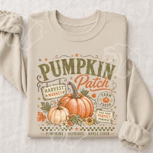 Könnte beinhalten: Ein sandfarbenes Sweatshirt mit Rundhalsausschnitt und einem Grafikdruck mit dem Text PUMPKIN PATCH, HARVEST MARKET, FARM FRESH und PICK YOUR PERFECT PUMPKIN. Das Design enthält Illustrationen von zwei orangefarbenen Kürbissen, Blättern und Blumen.