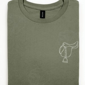 Könnte beinhalten: Ein olivgrünes T-Shirt mit Rundhalsausschnitt und einer weißen Strichzeichnung eines englischen Pferdesattels auf der rechten Seite. Das Shirt hat einen gerippten Kragen und ein schwarzes Gildan-Markenetikett an der Innenseite des Halsausschnitts.