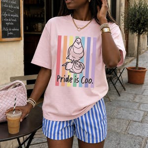 Könnte beinhalten: Hellrosa T-Shirt mit einer Grafik von zwei Tauben und dem Text "Pride is Coo". Das Design umfasst Regenbogenstreifen. Dazu blaue und weiße gestreifte Shorts. Auch goldener Schmuck ist zu sehen.