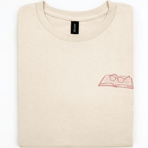Op de afbeelding: Een zandkleurig t-shirt met ronde hals en een kleine, rode lijntekening aan de rechteronderkant. De afbeelding toont een open boek met een bril op de pagina's. Het t-shirt heeft een zwart Gildan-merklabel bij de halslijn.