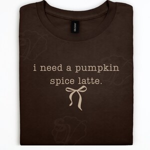 Op de afbeelding: Een donkerbruin t-shirt met de tekst "i need a pumpkin spice latte." in een lichtbeige kleur, vergezeld van een eenvoudige lijntekening van een strik onder de tekst. Het shirt heeft een ronde hals en een zwart merklabel in de nek.