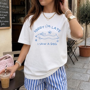 Könnte beinhalten: Ein weißes Kurzarm-T-Shirt mit einer blauen Grafik eines laufenden Dackels und dem Text Sorry I'm Late I Saw A Dog, kombiniert mit blau-weiß gestreiften Shorts. Das T-Shirt ist mit kleinen Sternen und Pfotenabdrücken verziert.