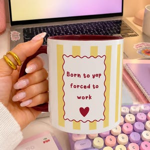 Könnte beinhalten: Ein weißer Keramikbecher mit vertikalen gelben Streifen sowie einem dunkelroten Innenraum und Henkel. Auf der Vorderseite befindet sich ein rechteckiger Rahmen mit gewelltem Rand und dem roten Text "Born to yap forced to work" über einem kleinen roten Herz.