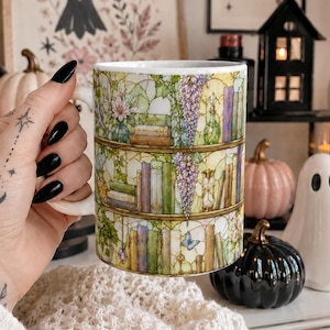Könnte beinhalten: Ein weißer Keramikbecher mit einer umlaufenden Illustration von drei Holzregalen, die mit bunten Büchern, hängenden Glyzinienranken, kleinen Topfpflanzen und einer Sanduhr gefüllt sind, vor einem Hintergrund aus Buntglas.