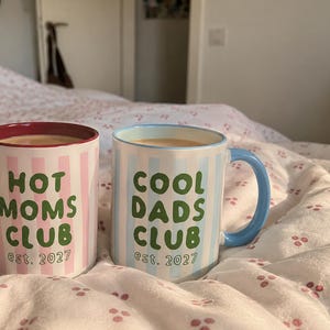 Peut inclure: Deux tasses en céramique à rayures verticales. La tasse de gauche est rose et blanche avec une anse rouge foncé et le texte vert HOT MOMS CLUB est. 2027. La tasse de droite est bleue et blanche avec une anse bleu clair et le texte vert COOL DADS CLUB est. 2027.