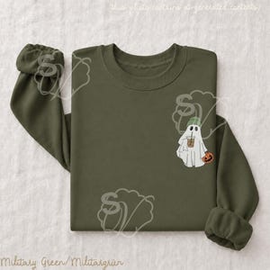 May include: An olive green crewneck sweatshirt featuring a small graphic on the left chest. The graphic depicts a white ghost wearing a green beanie, holding an iced coffee in one hand and a small orange jack-o'-lantern in the other hand.