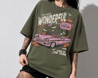 May include: An olive green t-shirt featuring a graphic print of a pink convertible car driven by skeletons. The design includes orange pumpkins, bats, and a sign reading THE MOST WONDERFUL HAUNTED BOOKSTORE SPOOKY SALE TIME OF THE YEAR in white text.