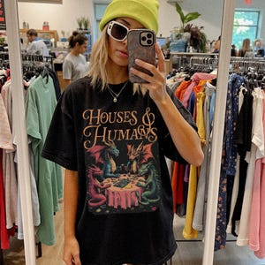 May include: Black t-shirt with the text "HOUSES & HUMANS" in a decorative font. The shirt features a colorful graphic of dragons around a table. The dragons are in shades of red, blue, and green.