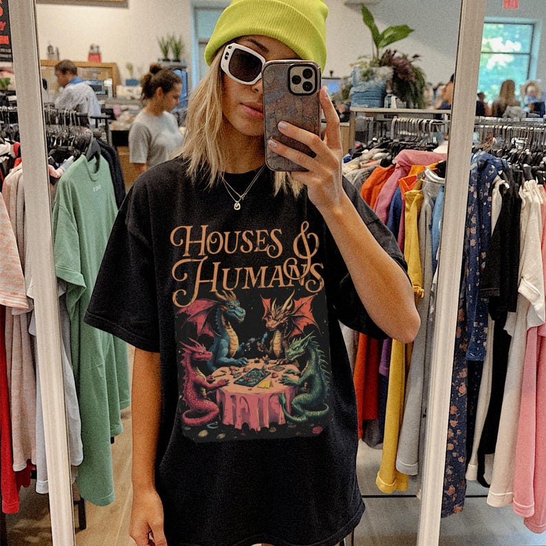 May include: Black t-shirt with the text "HOUSES & HUMANS" in a decorative font. The shirt features a colorful graphic of dragons around a table. The dragons are in shades of red, blue, and green.
