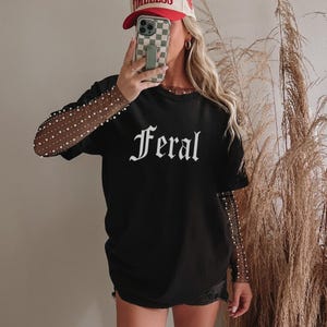 Könnte beinhalten: Schwarzes T-Shirt mit dem Wort "Feral" in weißen gotischen Buchstaben. Die Person trägt eine rot-beige Baseballkappe und ein durchsichtiges Langarmshirt mit Perlenakzenten. Das Outfit wird durch schwarze Shorts ergänzt.
