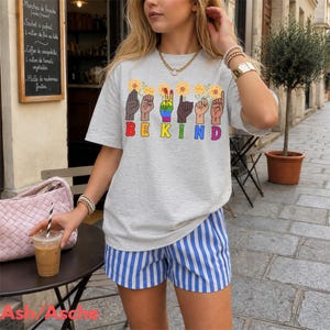 Könnte beinhalten: Ein hellgraues T-Shirt mit den Worten "BE KIND" in bunten Buchstaben. Das Shirt zeigt Handgesten und Sonnenblumen. Die Person trägt blau-weiß gestreifte Shorts.