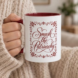 May include: A white ceramic mug with a dark red interior and handle. The front features a dark red floral border surrounding the text "Smash the Patriarchy" written in a decorative, flowing script font. The mug has a cylindrical shape.
