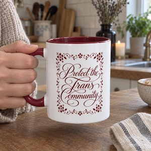 Könnte beinhalten: Ein weißer Keramikbecher mit dunkelrotem Innenraum und Henkel. Auf der Vorderseite steht der Text Protect the Trans Community in einer dekorativen Schreibschrift, umrahmt von einer rechteckigen Bordüre aus roten Blumenmustern.