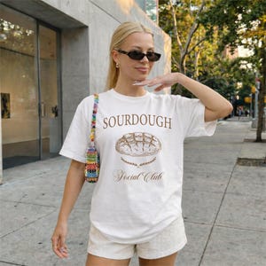 Op de afbeelding: Wit T-shirt met de woorden "SOURDOUGH Social Club" in bruin, en een broodafbeelding. Het shirt wordt gedragen met lichtgekleurde shorts en een kleurrijke kralentas.