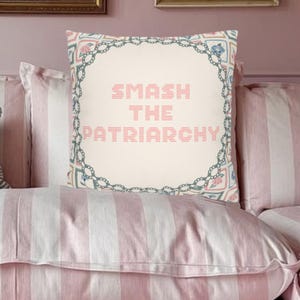 Könnte beinhalten: Ein dekoratives Kissen mit dem Text "SMASH THE PATRIARCHY" in rosa Kreuzstich. Das Kissen hat einen floralen und geometrischen Rand in Blau und Rosa, vor einem gestreiften rosa-weißen Hintergrund.