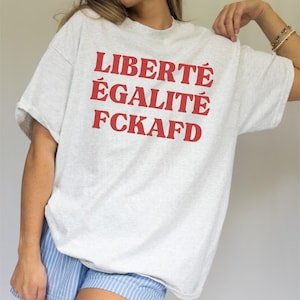 Könnte beinhalten: Hellgraues T-Shirt mit rotem Aufdruck "LIBERTÉ ÉGALITÉ FCKAFD". Das Shirt hat einen lässigen Stil mit kurzen Ärmeln.